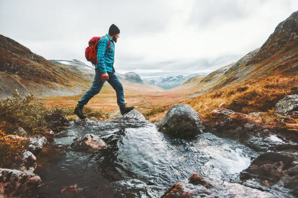 Man with a red backpack hiking across a stream in a mountain valley
